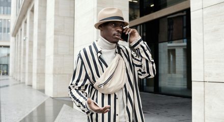 Fashionable man in a striped outfit and fedora engaging in a phone conversation outdoors.